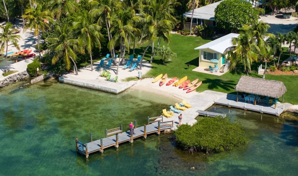 aerial view of bay harbor lodge in key largo there is a small beach area with kayaks, a small pier going out into the water with beach chairs, and multiple one story buildings in the background
