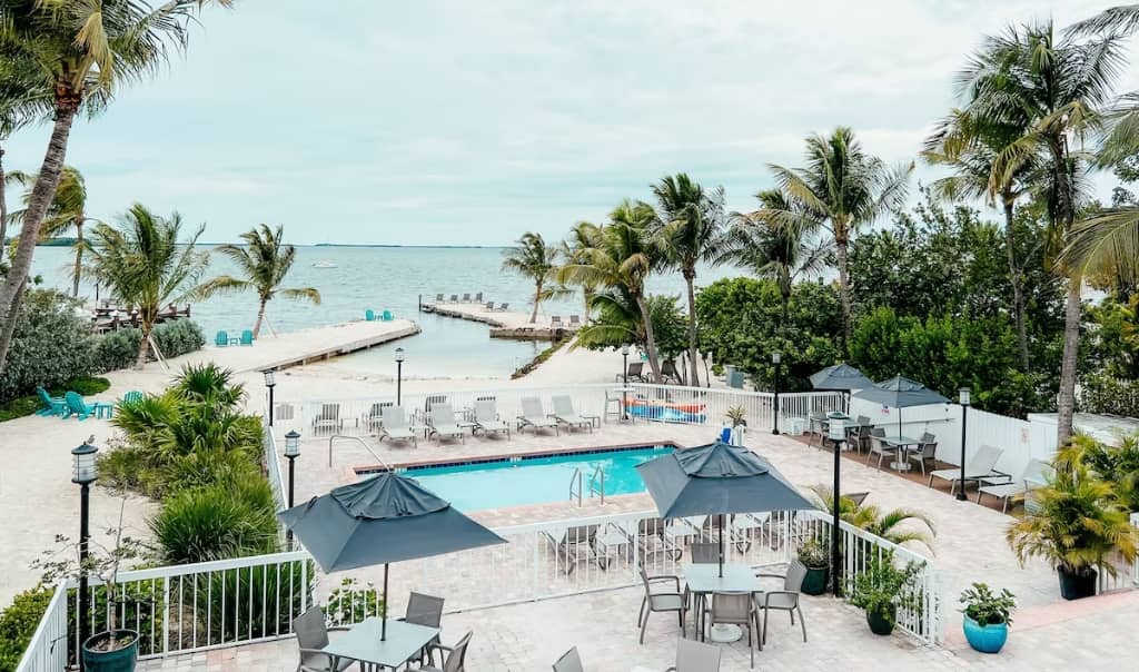 patio tables and chairs with umbrellas outside of a gated pool area with more sun chairs with a small beach in the background and a pier leading out to the beach and plam trees