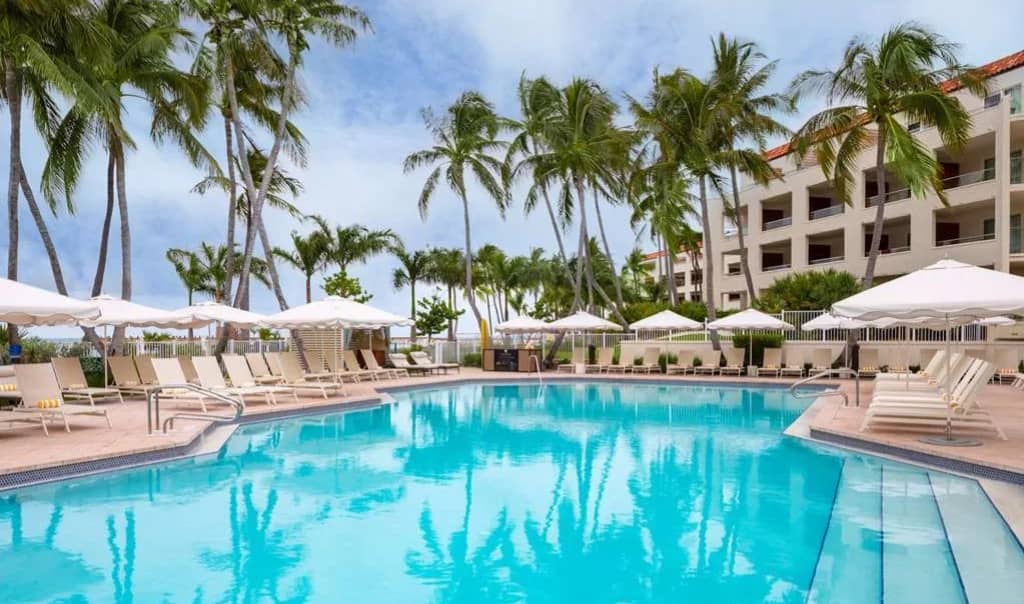 large pool surrounded by loungers with umbrella shades and palm trees with a three story resort in the background